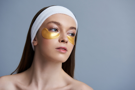 Close up portrait of attractive young woman with masks under lower eyelids looking away and keeping mouth slightly open. Isolated on blue backgroundの写真素材