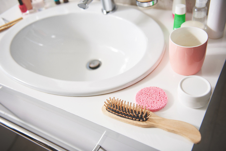 Self care and hygiene concept. Close up portrait of careful comb and cosmetic stuff on wash stand with white clean basinの写真素材