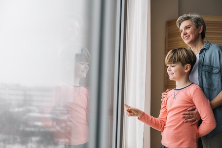Waist up photo of smiling granny and her grandson at window