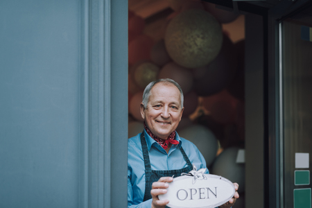 Joyful old man standing in the doorway of cafe and holding open sign