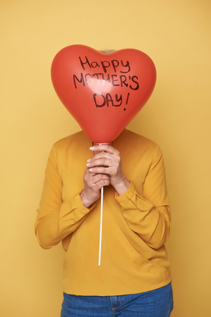 Stylish woman is hiding her head behind red balloon on yellow backgroundの写真素材