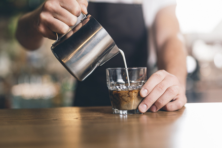 Barista pouring milk from the pitcher into glass of coffeeの写真素材