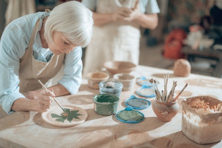 Cropped photo of elderly craftswoman painting decorative plate in workshopの写真素材