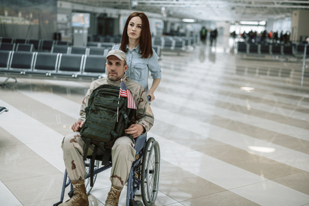 Wife carrying wheelchair and situating behind her husband in military uniformの写真素材