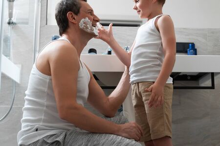Smiling dad and little son staying in bathroom and shavingの写真素材