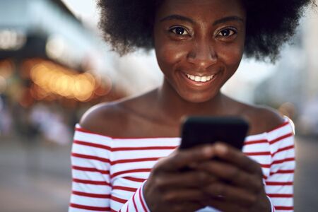 Happy afro-american lady is holding smartphone outdoorsの写真素材