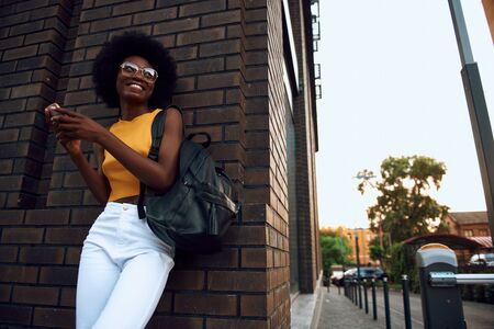 Joyful afro-american woman with bag holding smartphone outdoorsの写真素材