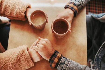 Love is in the air. Top view portrait of beloved man and woman holding hands and cups with hot beverage while sitting on table in cafeの写真素材