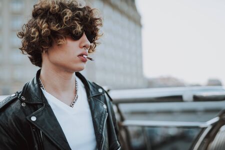 Curly rocker in sunglasses standing alone and smokingの写真素材