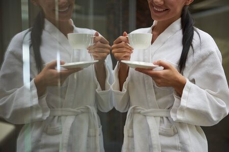 Joyful young lady with hot drink standing near glass wall with her reflectionの写真素材