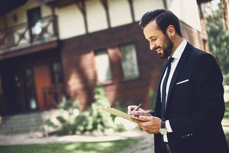 Cheerful estate agent standing near the house and making notesの写真素材