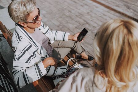 Smiling senior lady with a smartphone sitting with her friend on the benchの写真素材