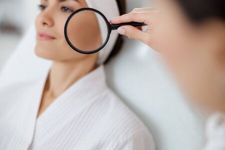 Cosmetologist holding loupe near face of smiling young woman stock photoの写真素材