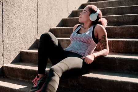 Charming young woman in headphones resting on stairs outdoorsの写真素材