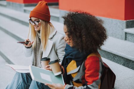 Young Caucasian and Afro american women sitting together with smartphoneの写真素材