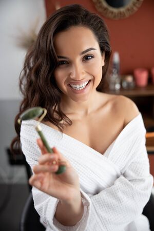 Cheerful young woman in bathrobe holding face massagerの写真素材