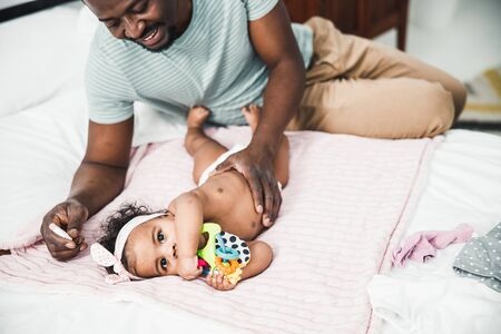 Cute Afro American baby girl lying on bed while smiling daddy placing hand on her belly stock photoの写真素材