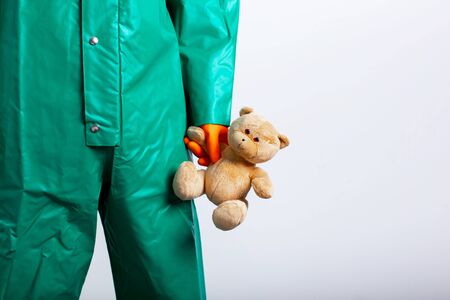 Cropped photo of a man in hazmat suit having orange rubber gloves on and holding a teddy bear....