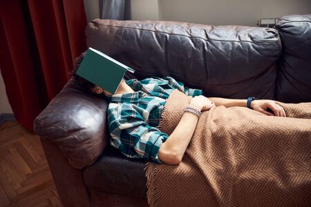Young man with book on face sleeping on sofa at homeの写真素材