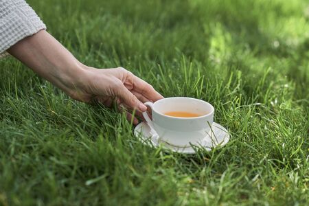 Close up of woman hand taking white porcelain mug of hot drink standing on grassの写真素材