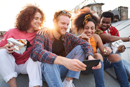 Cheerful friends with smartphone eating snacks on roof
