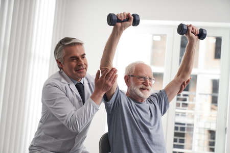 Smiling male rehabilitator helping elderly patient with exercisingの写真素材
