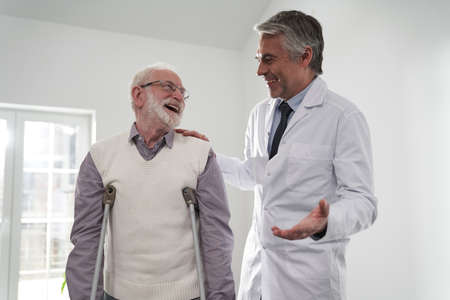 Happy aged man on crutches visiting cheerful physicianの写真素材