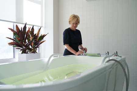 Worker of the spa salon preparing modern bathtub for health procedure