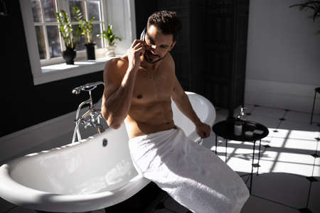 Handsome man wrapped in towel talking on mobile phone in bathの写真素材