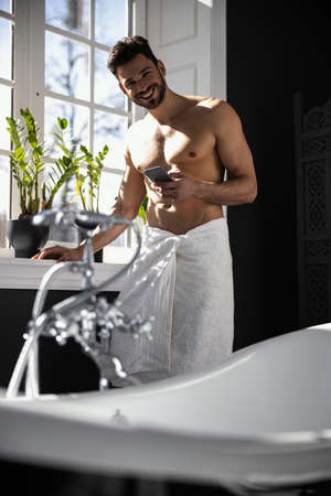 Handsome man wrapped in towel holding mobile phone in bathの写真素材