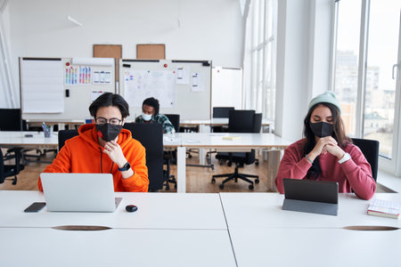Smart man wearing protective mask studying at designer faculty sitting at the desk near his group mateの写真素材