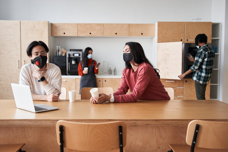 Two multiracial students wearing protective masks sitting at the table at the college kitchen