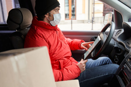 Delivery man in red uniform with protective mask at his face sitting at the car