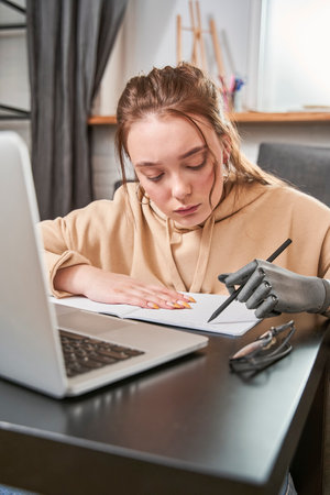Girl with prosthesis arm writing something at the notebook