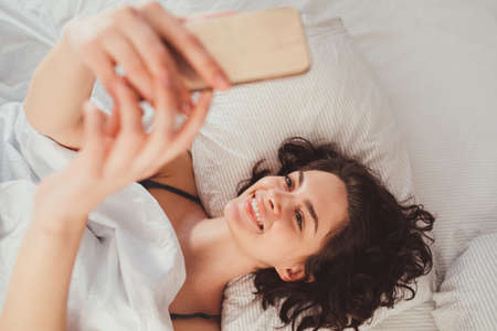 Beautiful woman using mobile phone to selfie while laying on bedの写真素材