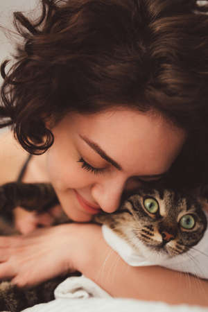 Young woman lying in bed and embracing her striped cat with green eyesの写真素材
