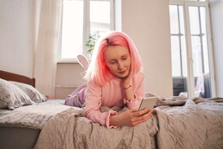 Woman with pink hair laying at the bed and typing something at her smartphoneの写真素材