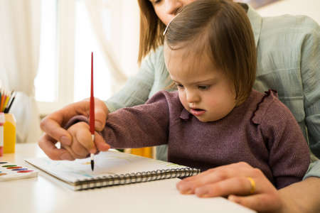 Kid with down syndrome drawing with paints on paper while sitting at the tableの写真素材