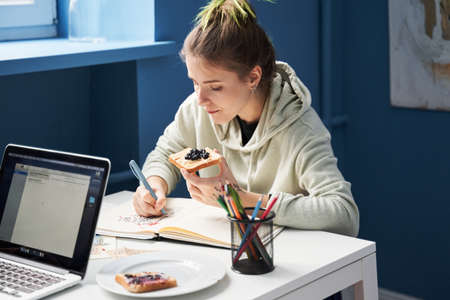 Woman sitting at the table and eating toast with jam while drawing sketches