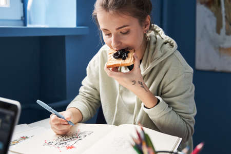 Woman sitting at the table and eating toast with jam while drawing sketchesの写真素材