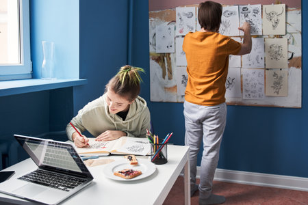 Man hanging tattoo sketches at the wall while his girlfriend drawing at the table