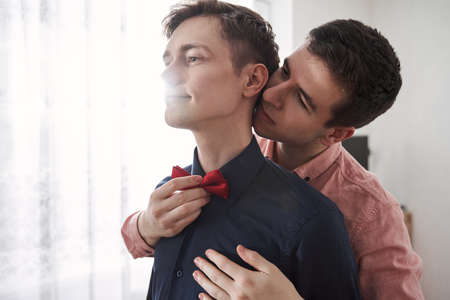 Gay man helping to put on a bowtie to his boyfriend while standing with him