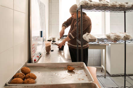 Baker standing at kitchen and carefully packing cakes with a scourge and paper