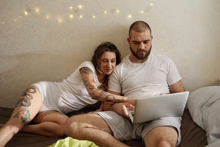 Woman looking at something at laptop while laying near her bearded boyfriend