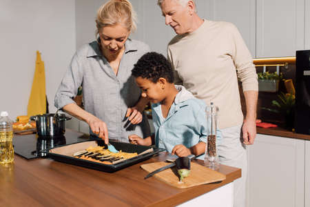 Caring grandparents teaching little boy cooking, while showing how greasing vegetablesの写真素材