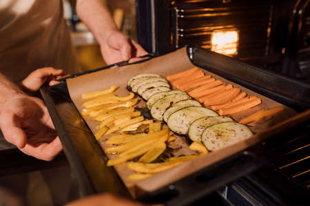 Senior man putting baking sheet with vegetables at the ovenの写真素材