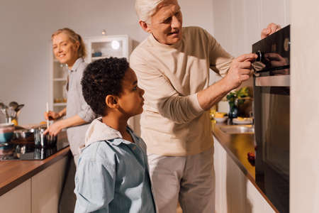 Grandfather and his multiracial grandson turning on oven while preparing dinnerの写真素材