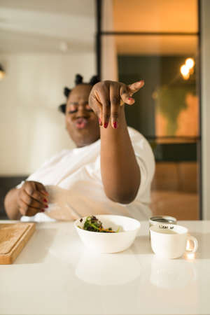 Woman putting salt into fresh vegetarian salad while sitting at the kitchenの写真素材