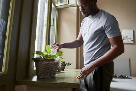 Man holding glass with water and watering flowers on the window at the morningの写真素材