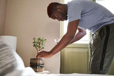 Man holding glass with water and carefully watering green plants at the nightstand near the bedの写真素材